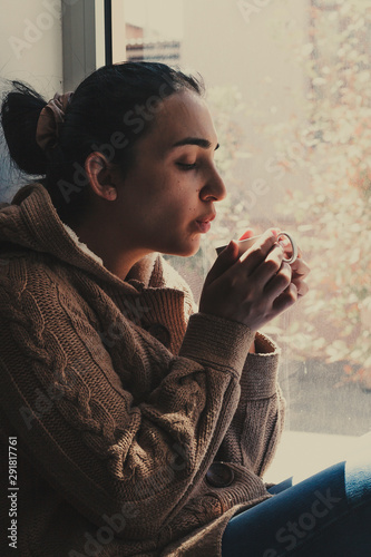 Young brunette woman drinking a cup of tea. Female sitting at home by the window. Cold autumn days. Fall background.