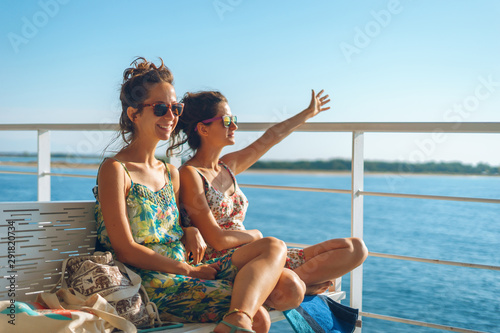 Two young women girl friends or sisters sitting on the bench on the deck of the ferry boat or ship sailing to the island tourist destination on summer vacation waving to the horizon in sunny day © Miljan Živković