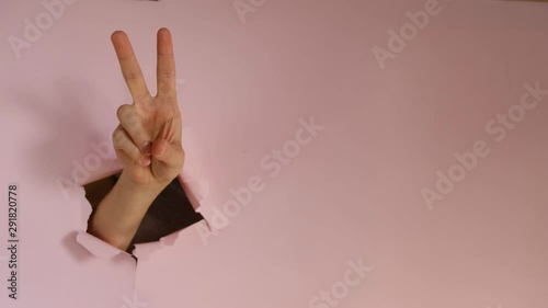 Closeup of female hand demonstrates victroy sign through torn hole in pink paper, expresses celebration and triumph. Body language concept. Hand sign.  Breaking through paper wall.  photo