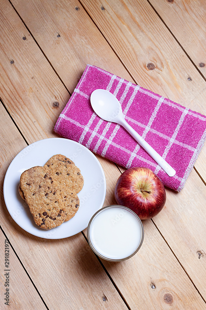 TOP VIEW OF BREAKFAST WITH COOKIES WITH CHOCOLATE CHIPS, MILK GLASS AND RED APPLE WITH KITCHEN CLOTH AND SPOON ON WOODEN TABLE. HEALTHY BREAKFAST TOP VIEW OF BREAKFAST WITH COOKIES WITH CHOCOLATE CHIPS, MILK GLASS AND RED APPLE WITH KITCHEN CLOTH AND SPOON ON WOODEN TABLE. HEALTHY BREAKFAST