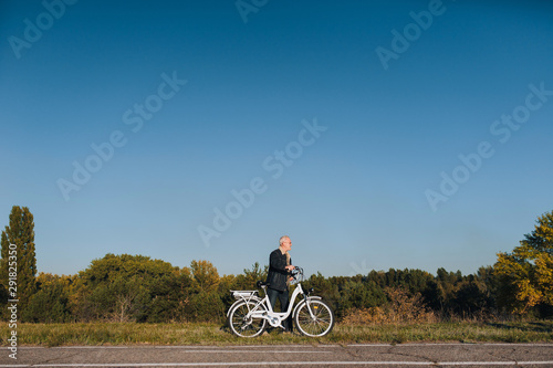 Pensioner. An elderly man stand near an electric white bike on an asphalt road against the background of autumn nature. The concept of a happy old age and pension.