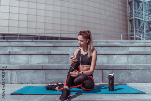 A girl in the summer in the city at a workout, resting after a workout on a yoga mat, looking at the smartphone application on a timer on the phone, a shaker with protein and water.