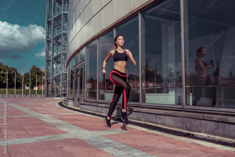 Stylish fashionable slender woman. Beautiful athletic tanned athlete girl run summer city background glass windows, free space for fitness motivation text. Sportswear. Leggings. Top