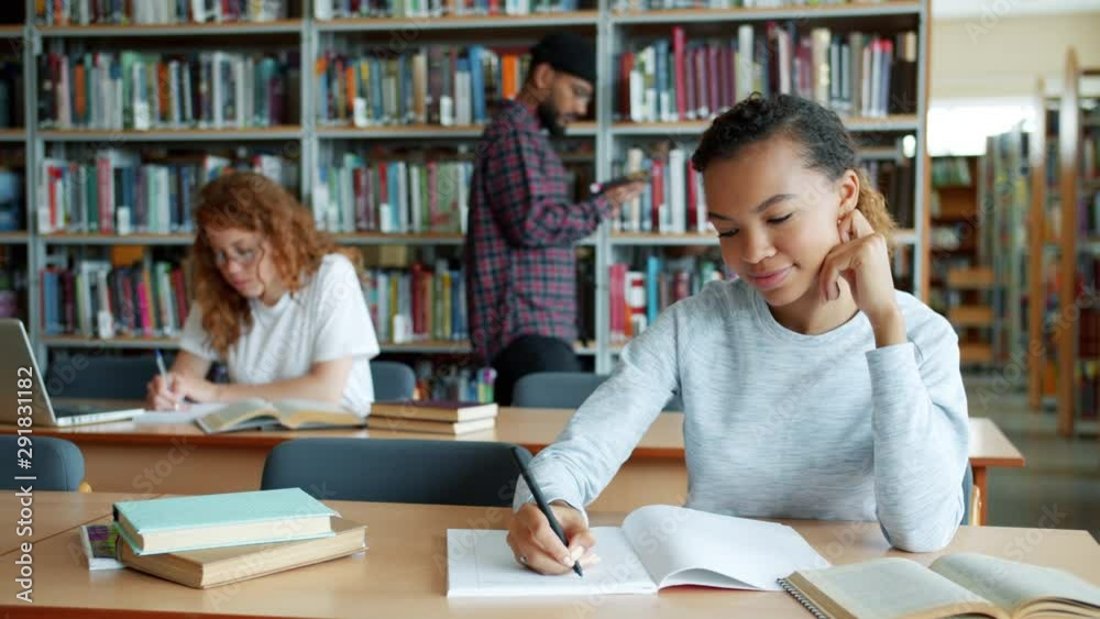 African American girl writing reading studying in library while people are walking around book shelves in background. Education, lifestyle and knowledge concept. African American girl writing reading studying in library while people are walking around book shelves in background. Education, lifestyle and knowledge concept.