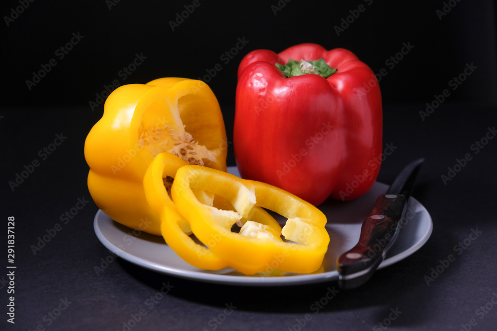 A sliced yellow bell pepper and whole red bell pepper together on small plate with sharp knife on dark background A sliced yellow bell pepper and whole red bell pepper together on small plate with sharp knife on dark background