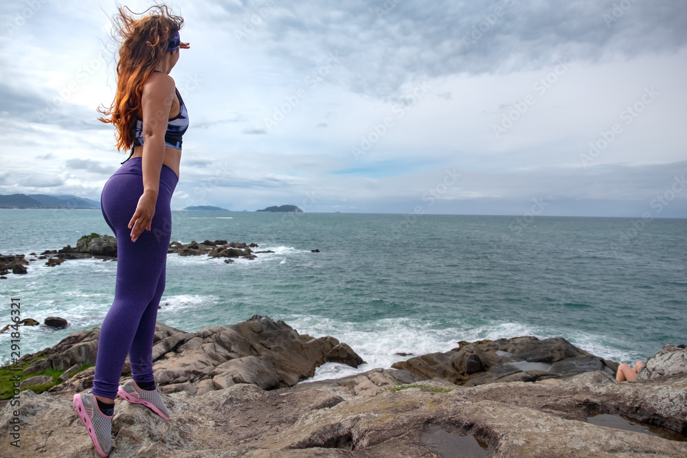 Mujer con ropa deportiva  de pie sobre rocas observando un mar azul en un día soleado