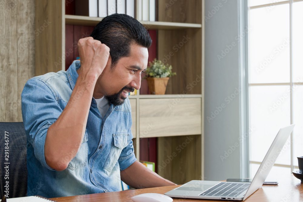 Handsome bearded guy is sitting at the desk, He raise fisting expressing confidence after successful work. Asian online business entrepreneur is shouted gladly when a customer orders the product. Handsome bearded guy is sitting at the desk, He raise fisting expressing confidence after successful work. Asian online business entrepreneur is shouted gladly when a customer orders the product.