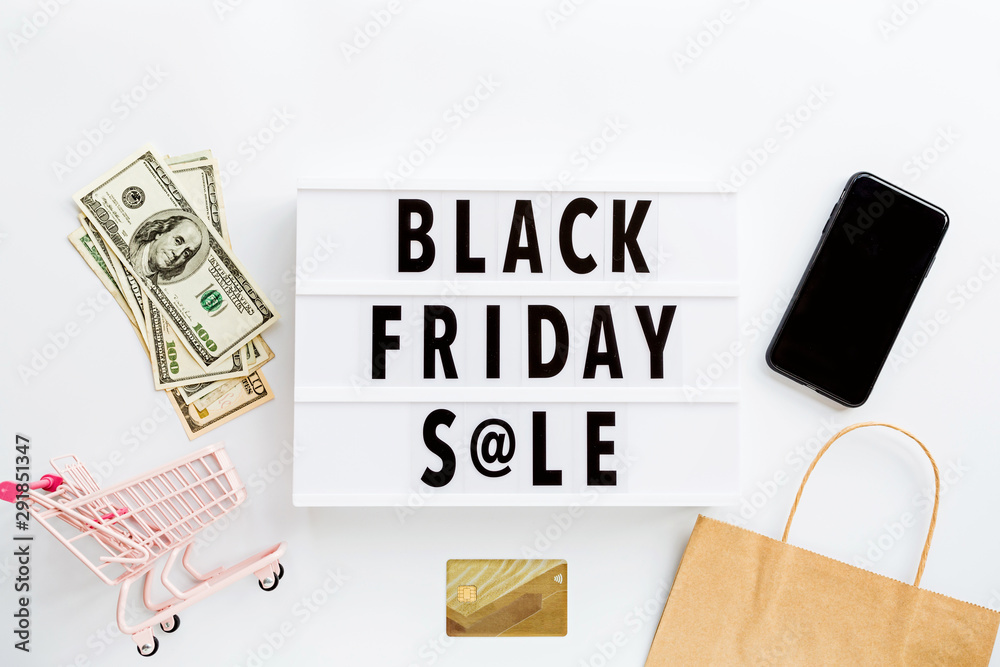 Creative promotion composition Black friday sale text on lightbox on white background, next grocery trolley, credit card, cash money, mobile phone, shopping bag. Flat lay, top view, overhead, mockup