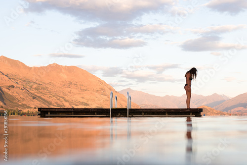 A lone girl contemplates swimming at dawn in a still lake, surrounded by mountains photo