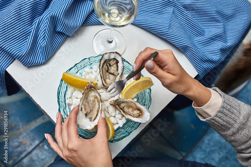 The girl's hands hold a fork and an open fresh oyster on the background of plates with lemon slices, oysters and a glass of wine on the table with a blue tablecloth. Dinner at the restaurant photo