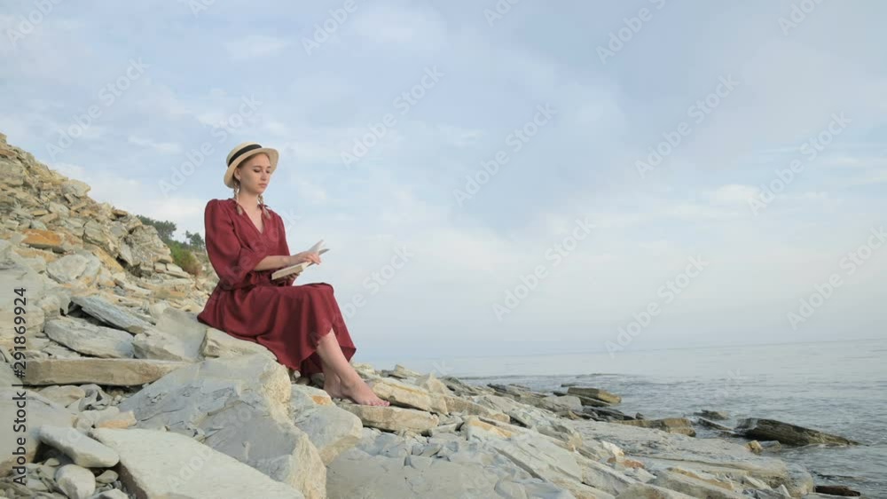 An attractive Caucasian girl in a red bright dress and a straw hat sits on a large stone by the sea at sunset and reads an interesting book on the sea horizon and beautiful clouds. An attractive Caucasian girl in a red bright dress and a straw hat sits on a large stone by the sea at sunset and reads an interesting book on the sea horizon and beautiful clouds.