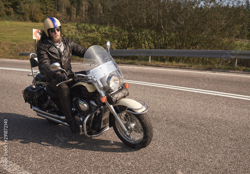 Bearded motorcyclist in helmet, sunglasses and black leather clothing riding cruiser motorbike along sharp turn of empty road on bright summer day