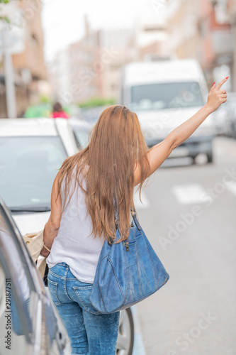 Rear view of blonde girl raising her hand as if she wanted to stop a taxi wearing white shirt with blue pants, blue wallet with blurred background. Vertical image.