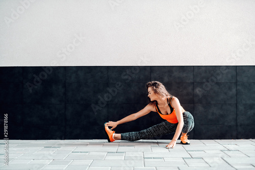 Beautiful slim caucasian sporty woman in sportswear and with curly hair stretching her leg before running. In background is black and white wall.