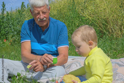 Grandfather and cute blond grandson are eating and sitting on a picnic together in summer park outdoor. Authentic lifestyle image. Fatherhood and childhood and generation concept. © Nataliia