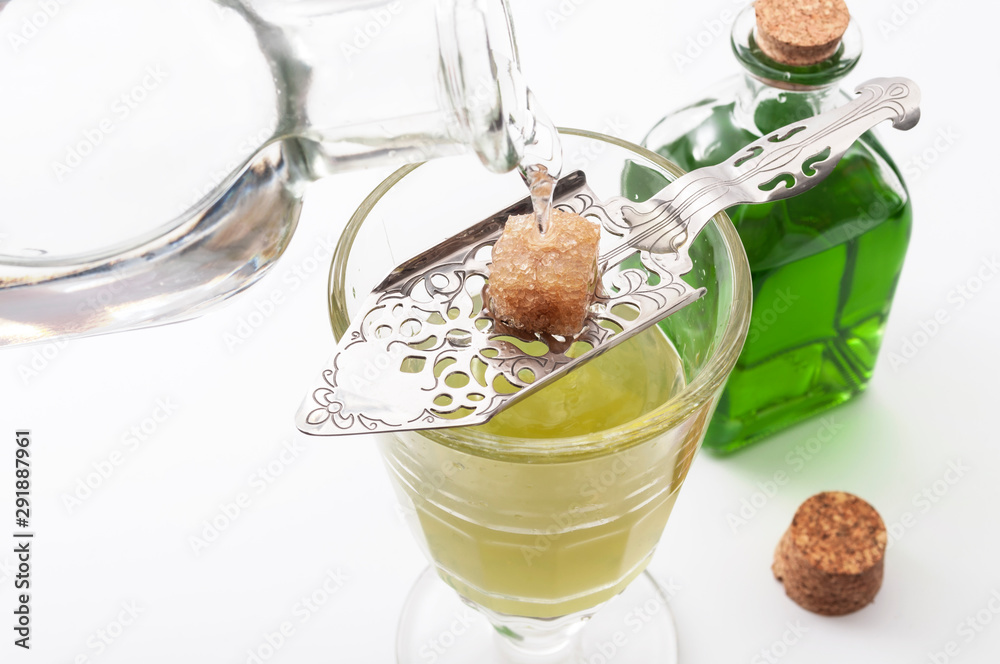 Alcohol, luxurious spirits and dangerous liquor conceptual idea with crystal glass of absinthe, pouring water on stainless steel spoon, sugar cube and green bottle of strong spirit on white background