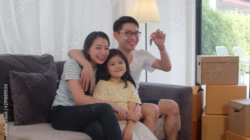 Portrait of Happy Asian young family bought new house. Japanese little preschool daughter with parents mother and father holds in hand keys sitting on sofa in living room smiling looking at camera.