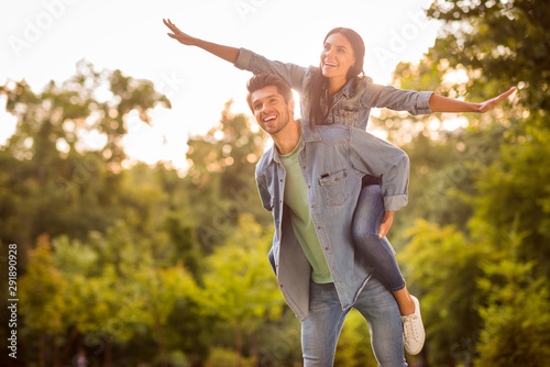 Portrait of cheerful man and his woman raising her hands piggyback having brunette hair wearing denim jeans outdoors