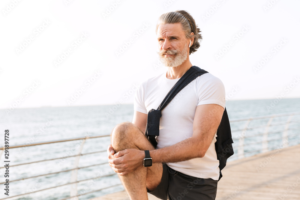 Image of handsome old man using earpod while stretching his leg