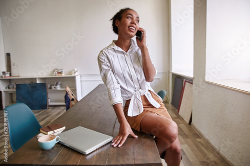 Pretty young dark skinned female with casaul hairsyle wearing striped white shirt, taking break from work and making call to friend, sitting on table photo