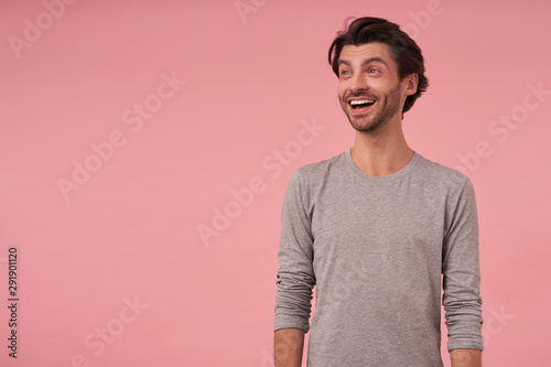 Studio postrait of joyful handsome dark haired male with beard wearing grey sweater, standing over pink background, looking aside with broad smile, contracting forehead and raising eyebrows