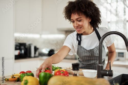 Beautiful mixed race housewife in apron reaching for tomatoes while standing in kitchen. On kitchen counter are vegetables. Dinner preparation.