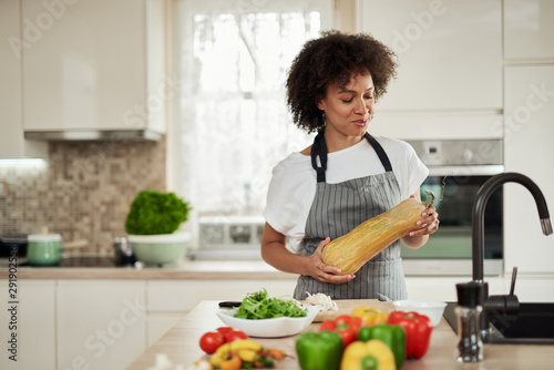 Beautiful mixed race housewife in apron standing in kitchen and holding zucchini On kitchen counter are vegetables. Meal preparation.