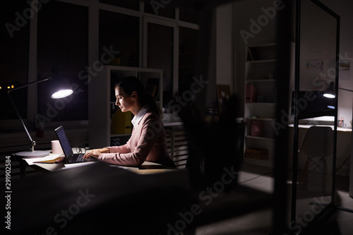 Young manager sitting at her workplace and typing on laptop computer under the light of lamp she working in dark office till night