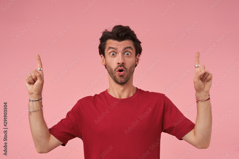 Surprised young dark haired male with beard standing over pink background in red t-shirt, looking at camera amazedly and pointing upwards with index fingers, contracting forehead and raising eyebrows Surprised young dark haired male with beard standing over pink background in red t-shirt, looking at camera amazedly and pointing upwards with index fingers, contracting forehead and raising eyebrows