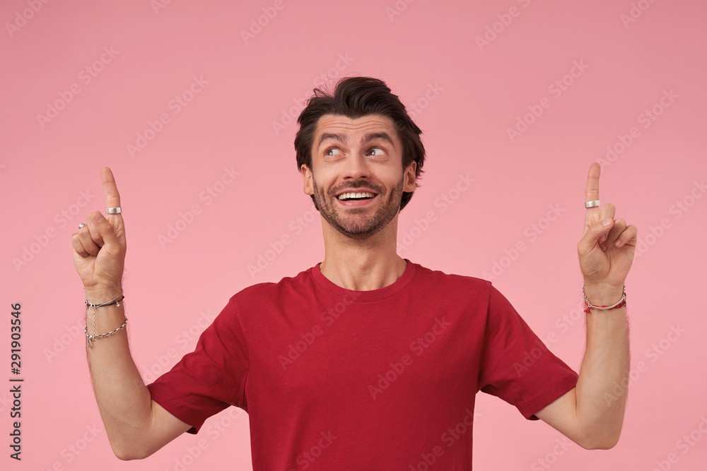 Happy attractive bearded male wearing red t-shirt, looking upwards and pointing up with forefingers, standing over pink background, contracting forehead and raising eyebrows with wide smile