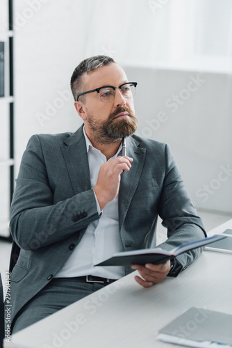 handsome businessman in formal wear and glasses holding pen and notebook in office