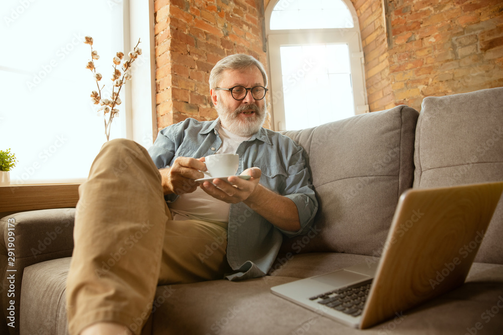 Senior man working with laptop at home - concept of home studying. Caucasian male model sitting on sofa and having coffee break while serfing in internet, watching cinema or webinar, online lessons.