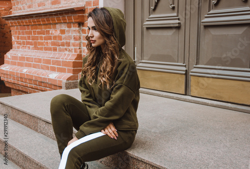 Fototapeta Naklejka Na Ścianę i Meble -  young casual girl with long brunette hair in khaki sports costume with hood is sitting and looking straight on stairs near street's door and brick wall background, sport lifestyle concept, free space