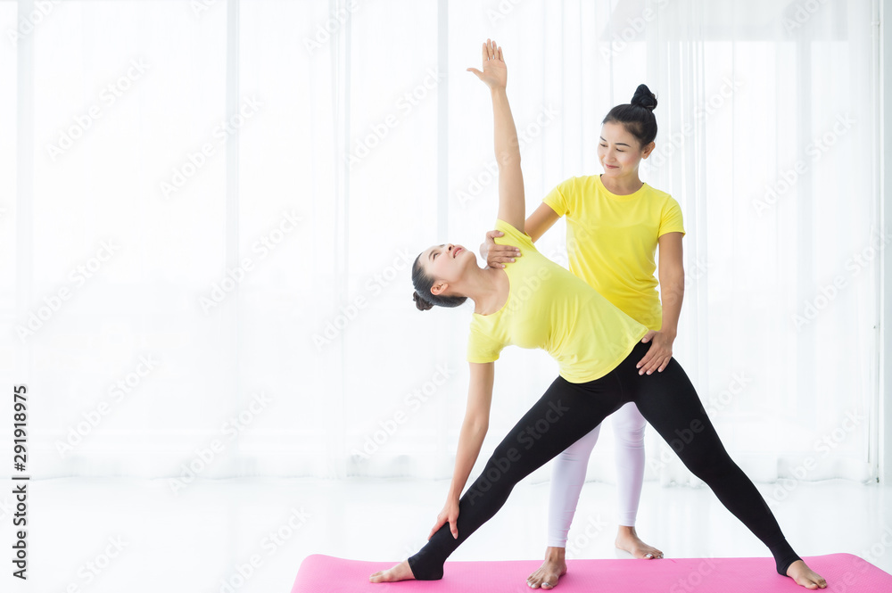 Two young Asian women workout practicing yoga in yellow dress or pose with a trainer and practice meditation wellness lifestyle and health fitness concept in a gym. Two young Asian women workout practicing yoga in yellow dress or pose with a trainer and practice meditation wellness lifestyle and health fitness concept in a gym.