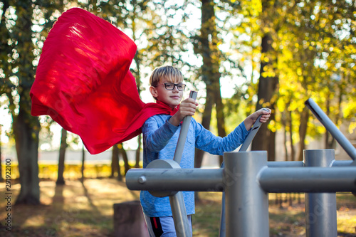 Fototapeta Naklejka Na Ścianę i Meble -  Smart boy with red cloak does workout on trainer equipment outdoor and feels himself like a hero. Children, sport, activity and healthcare concept