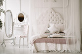White bedroom interior with nobody. large cozy bed with a white canopy and oval dressing mirror with dressing table next to it