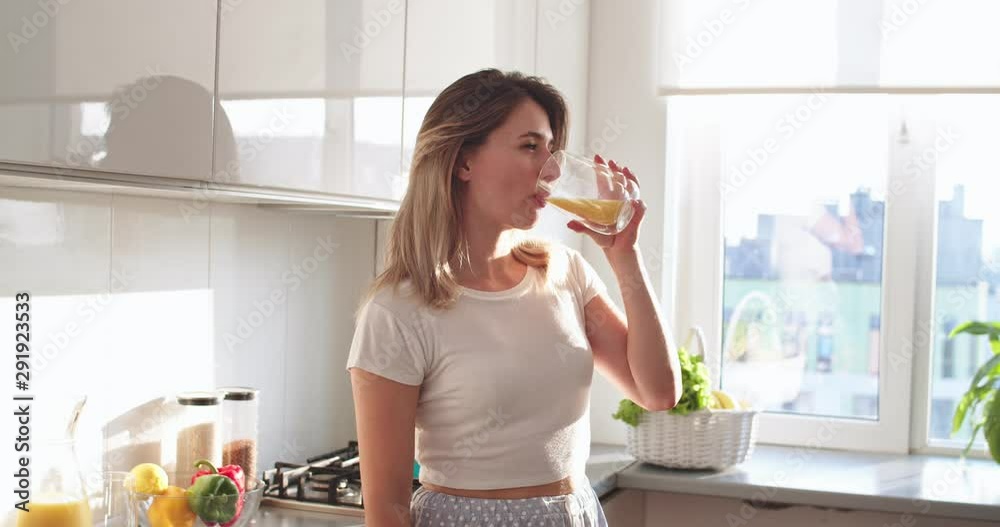 Portrait shot of the beautiful young Caucasian woman in the white T-shirt drinking orange juice early in the morning in the kitchen and then smiling cheerfully to the camera.