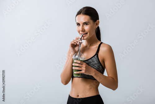 attractive smiling sportswoman drinking smoothie isolated on grey