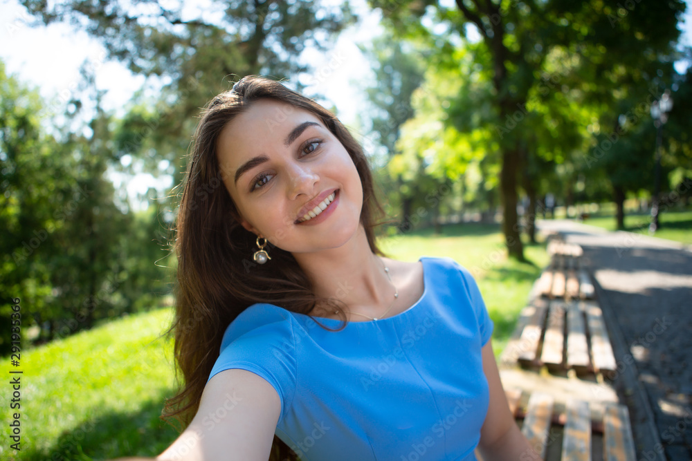 Happy young woman in a blue dress sits in a park and making selfie with the smartphone.