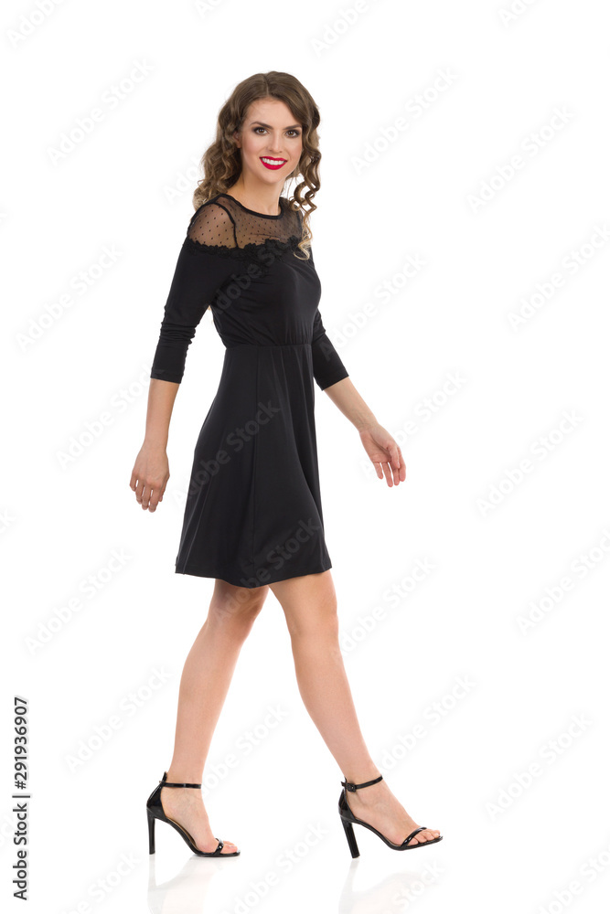 Young Woman In Elegant Black Dress And High Heels Is Walking And Looking At Camera. Side View. Young Woman In Elegant Black Dress And High Heels Is Walking And Looking At Camera. Side View.