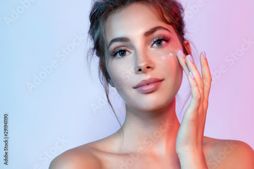 Cute woman with natural make-up applying moisturizing facial cream. Portrait of wonderful female looking at camera with calmness. Beauty and skincare concept