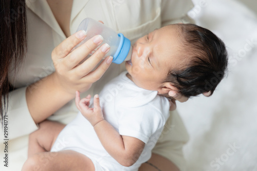 Young Asian mother hugging a cute newborn baby and feeding milk form bottle in white bedroom. Duty of the wife to raise children. Mother and child concept
