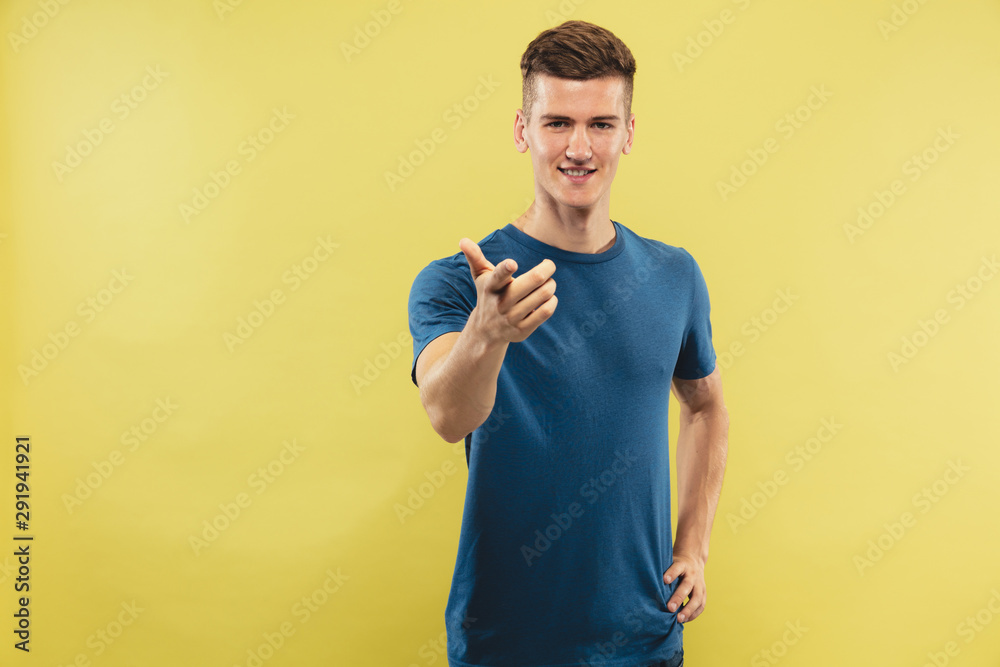 Caucasian young man's half-length portrait on yellow studio background. Beautiful male model in blue shirt. Concept of human emotions, facial expression. Pointing on and smiling, choosing and inviting Caucasian young man's half-length portrait on yellow studio background. Beautiful male model in blue shirt. Concept of human emotions, facial expression. Pointing on and smiling, choosing and inviting