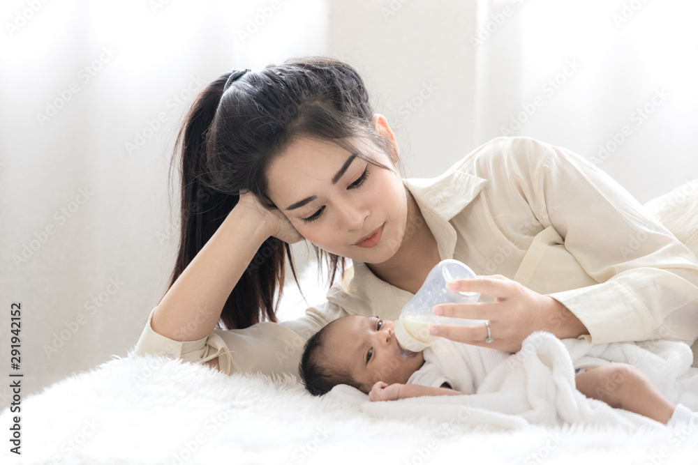 Young Asian mother hugging a cute newborn baby and feeding milk form bottle in white bedroom. Mother and child concept Young Asian mother hugging a cute newborn baby and feeding milk form bottle in white bedroom. Mother and child concept