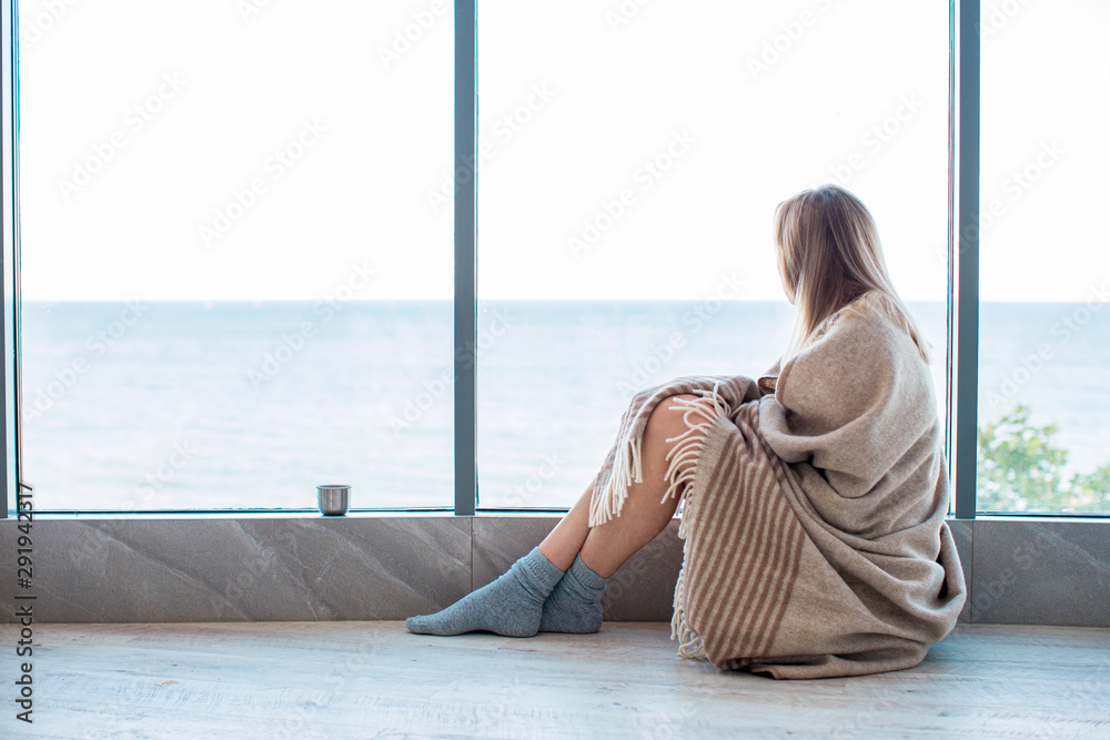 Relaxed Caucasian woman sitting on a warm floor in a socks wrapped in a woolen blanket near the large window in light. autumn mood, warmth and comfort.