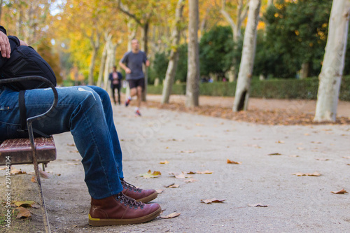 A man  who is spending the time spending time outdoors and observing the people that is walking in the street while he is sitting in a bench of a park in fall season photo