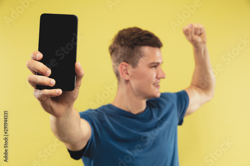 Caucasian young man's half-length portrait on yellow studio background. Beautiful male model in blue shirt. Concept of human emotions, facial expression. Showing phone's screen and looks celebrating.