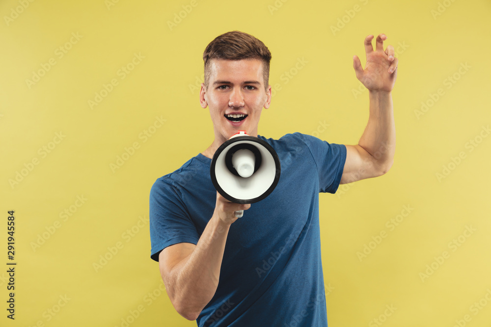 Caucasian young man's half-length portrait on yellow studio background. Beautiful male model in blue shirt. Concept of human emotions, facial expression, sales. Calling, screaming with mouthpeace. Caucasian young man's half-length portrait on yellow studio background. Beautiful male model in blue shirt. Concept of human emotions, facial expression, sales. Calling, screaming with mouthpeace.