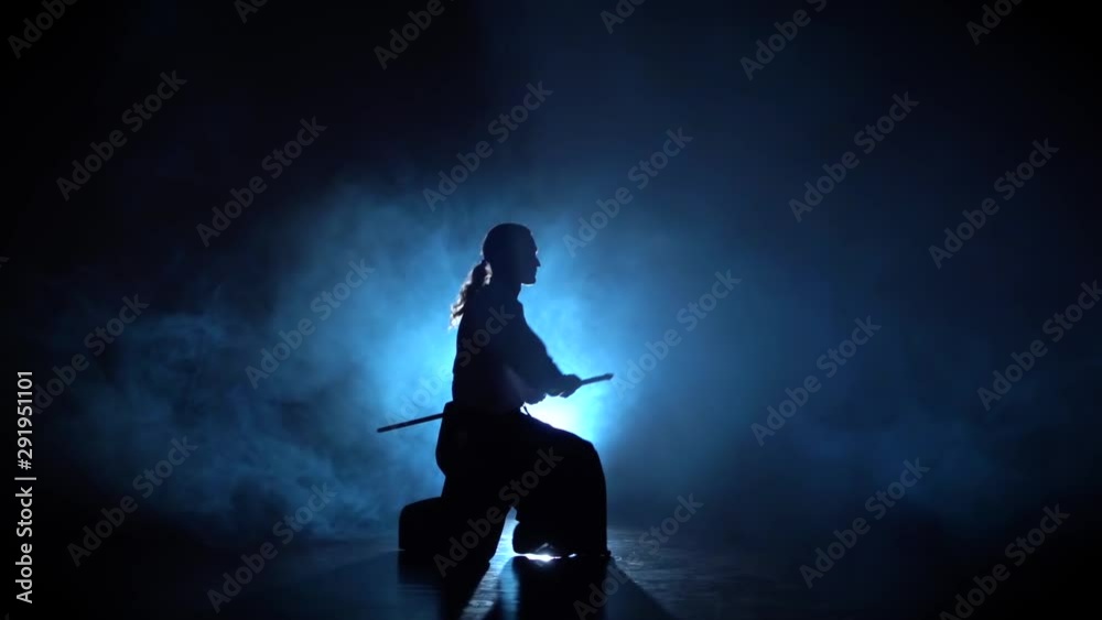 Silhouette Aikido Master Wearing Traditional Samurai Hakama Clothes Takes His Japanese Sword out of Scabbard and Swings with It, from a sitting position. He's in the Spotlight Darkness Surrounds Him. Silhouette Aikido Master Wearing Traditional Samurai Hakama Clothes Takes His Japanese Sword out of Scabbard and Swings with It, from a sitting position. He's in the Spotlight Darkness Surrounds Him.