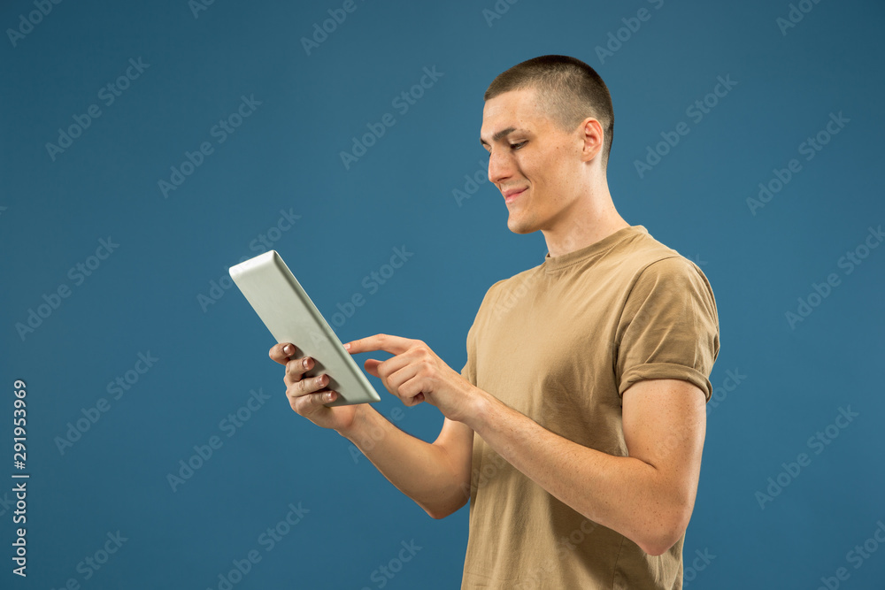 Caucasian young man's half-length portrait on blue studio background. Beautiful male model in shirt. Concept of human emotions, facial expression, sales, ad. Using tablet for selfie, vlog or study.
